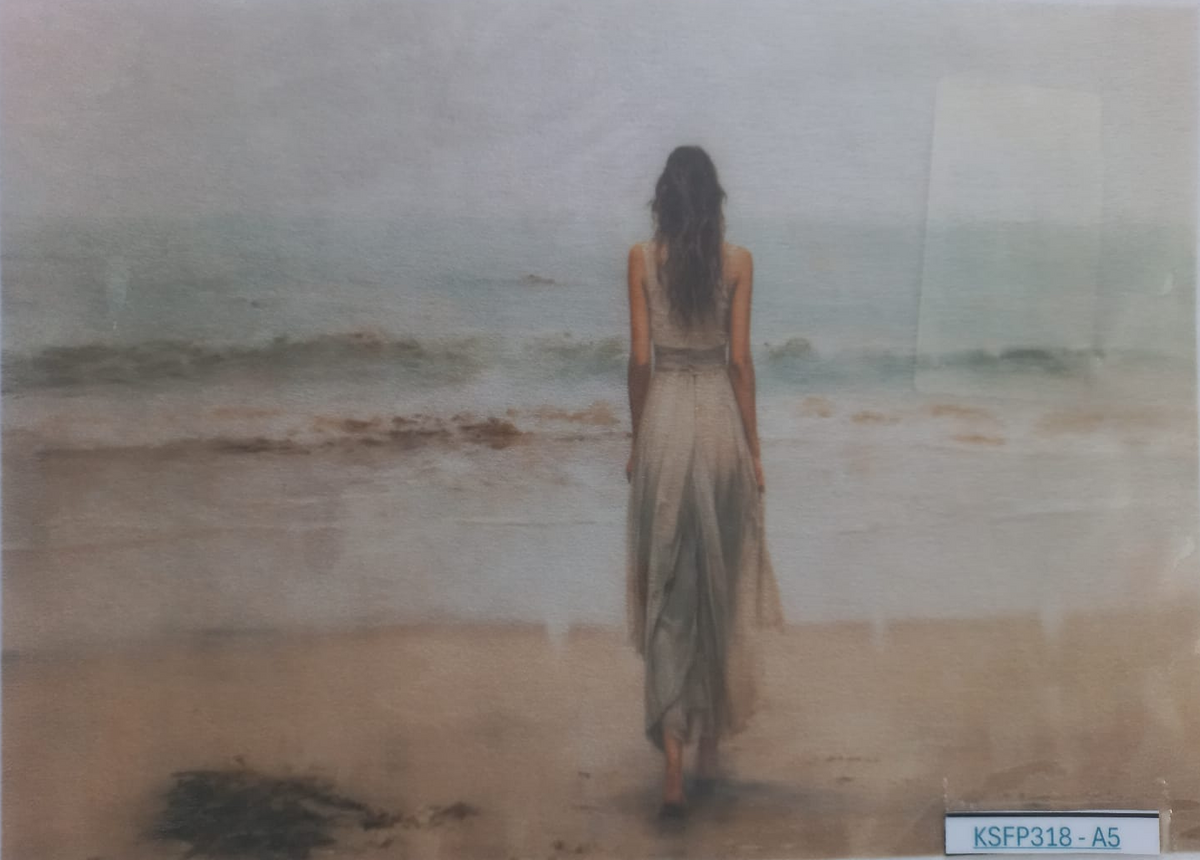 A photographic art print depicting a woman from behind walking onto a beach towards the ocean, mounted on a backing card with a white label at the bottom right.