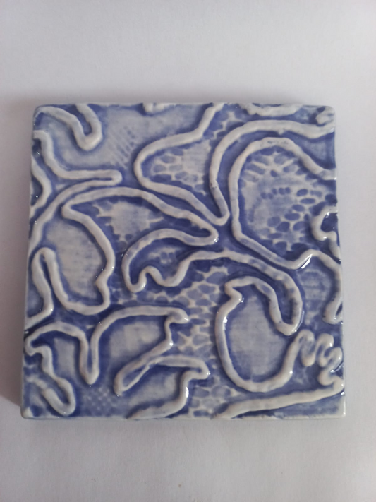 A square ceramic tile featuring an abstract, raised wavy pattern in cobalt blue and white glaze, photographed from directly above on a neutral light gray background.
