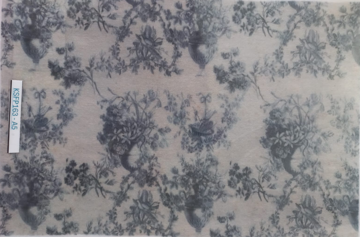 A square fabric sample with a classic toile de Jouy pattern in slate blue on a warm beige background, enclosed in a clear plastic sleeve with a white label on the left side.