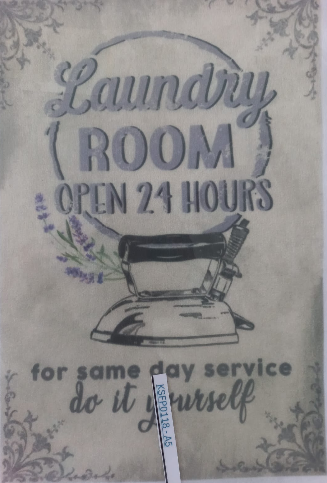 A vintage-style laundry room sign with floral borders, an illustration of an iron, and the text 'Laundry ROOM OPEN 24 HOURS for same day service do it yourself.' A small vertical label is attached near the bottom.