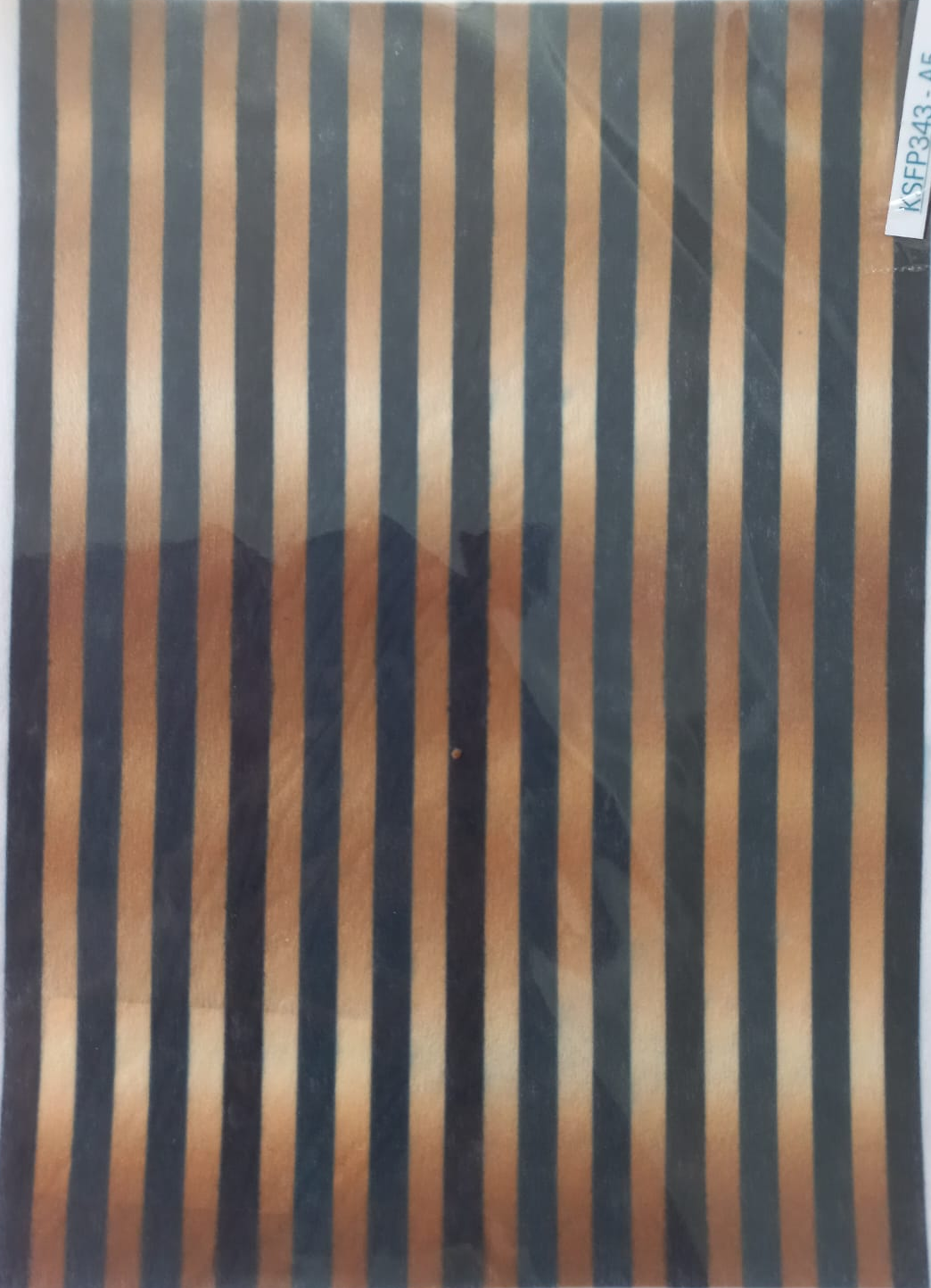 A sheet of patterned fabric or material inside a clear plastic sleeve, featuring vertical stripes in black, dark brown, and beige with a gradient effect.